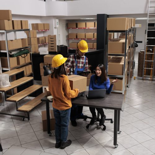 Warehouse package handlers doing inventory control while coworking with logistics manager. Asian storehouse employees scanning cardboard box and creating invoice on laptop